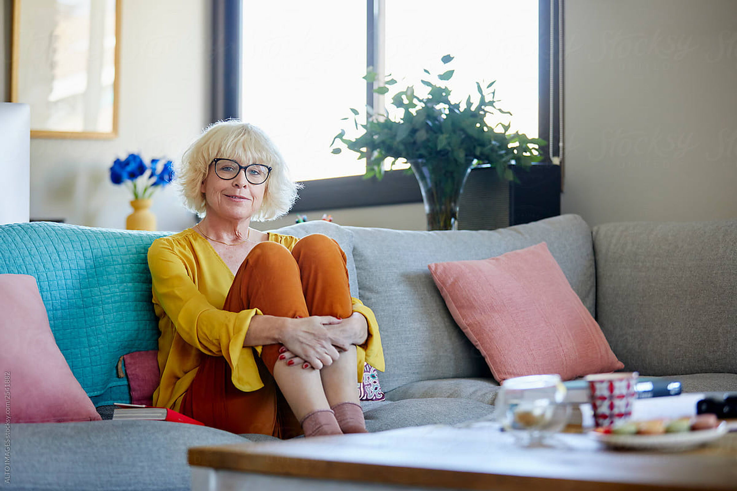 woman with glasses and bright clothes sitting on couch hugging her legs to her chest
