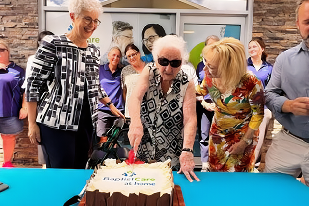 People surrounding BaptistCare cake and elderly woman cutting