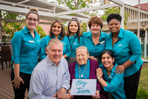 an elderly nursing home resident with a team of aged care workers holding a MyTeam poster at baptistcare niola centre aged care home in parkes nsw
