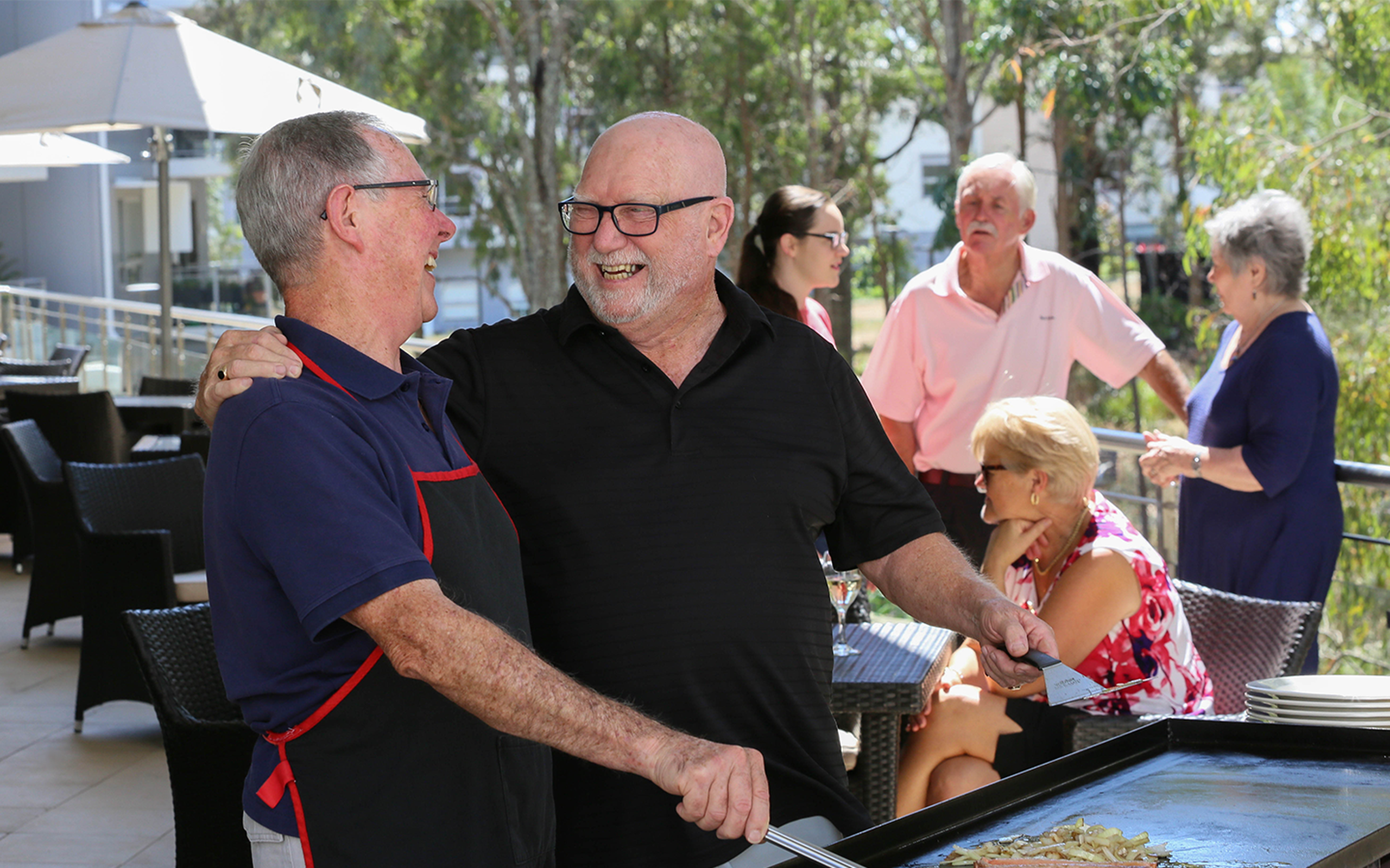 two over 55s men enjoying independent living in baptistcare retirement village cooking a bbq on the balcony socialising with their friends during their retirement